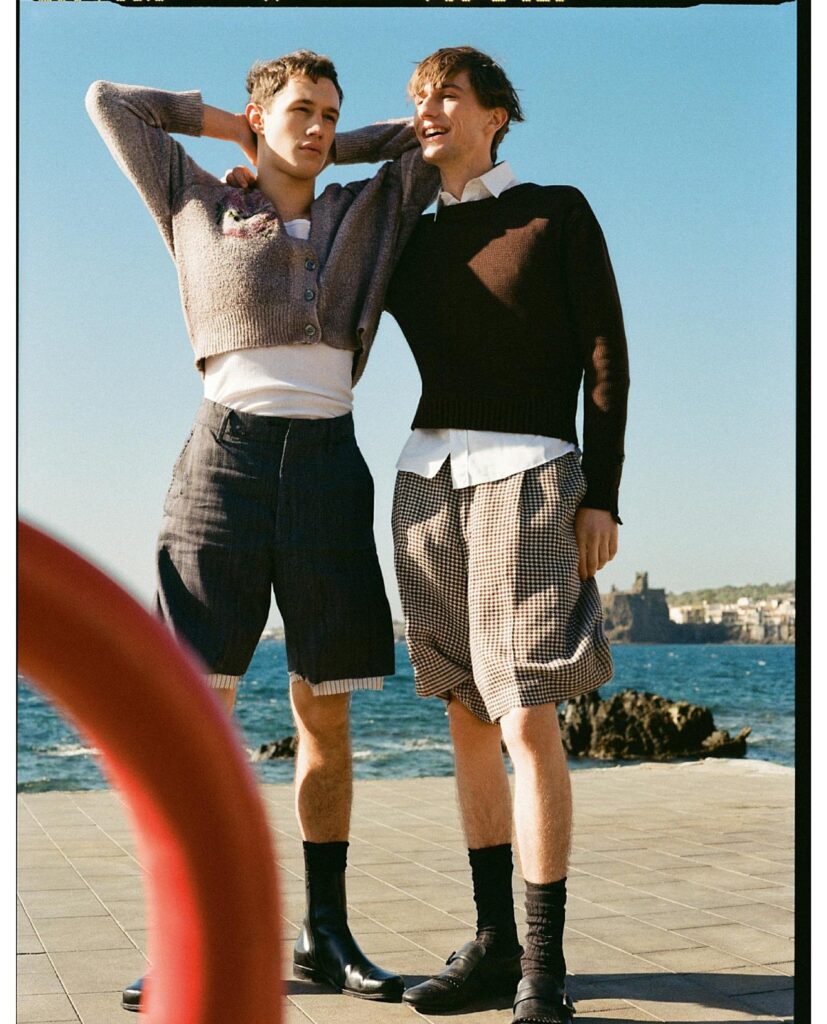 Two men standing with arms around each other by the seaside, wearing knit sweaters and shorts on a sunny day.