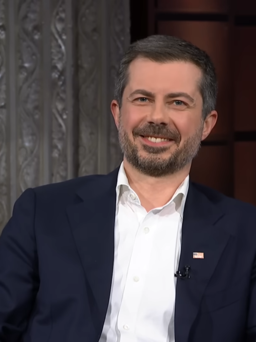 Smiling man in a dark suit and white shirt sits on a blue couch during a televised interview on a TV talk show set.