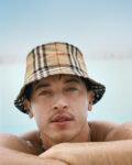 Close-up of a man in a beige plaid bucket hat resting at the edge of a pool with blue water and sky behind him.