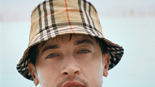 Close-up of a man in a beige plaid bucket hat resting at the edge of a pool with blue water and sky behind him.