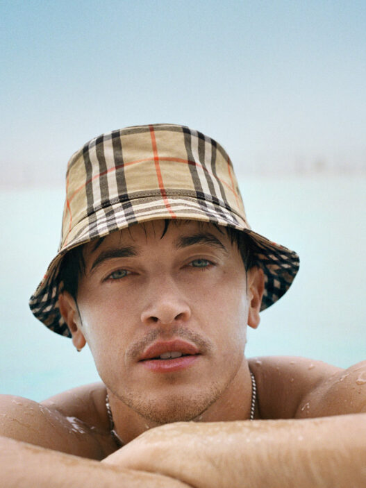 Close-up of a man in a beige plaid bucket hat resting at the edge of a pool with blue water and sky behind him.