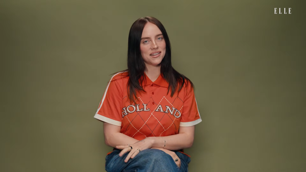 Portrait of a woman with long dark hair, wearing an orange shirt with a diamond pattern and the word 'HOLLAND,' sitting against an olive wall as she speaks to the camera.
