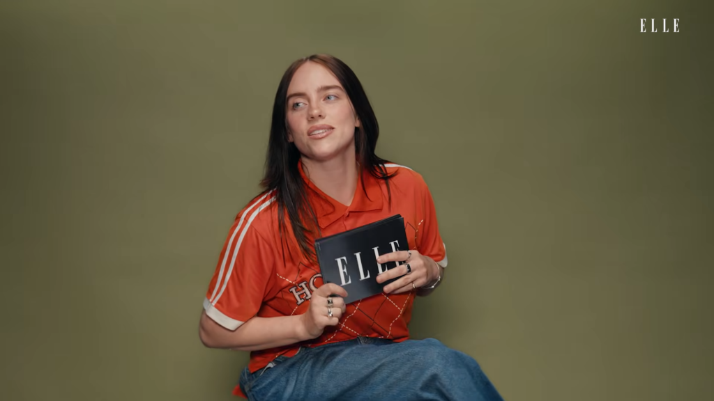 Woman with black hair in a red polo shirt holding an ELLE magazine against an olive-green backdrop.