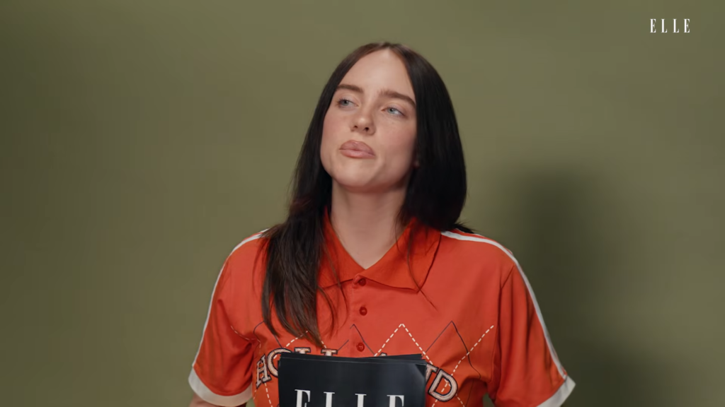 Woman with long dark hair in an orange polo speaking, ELLE logo in the top-right corner of the frame.