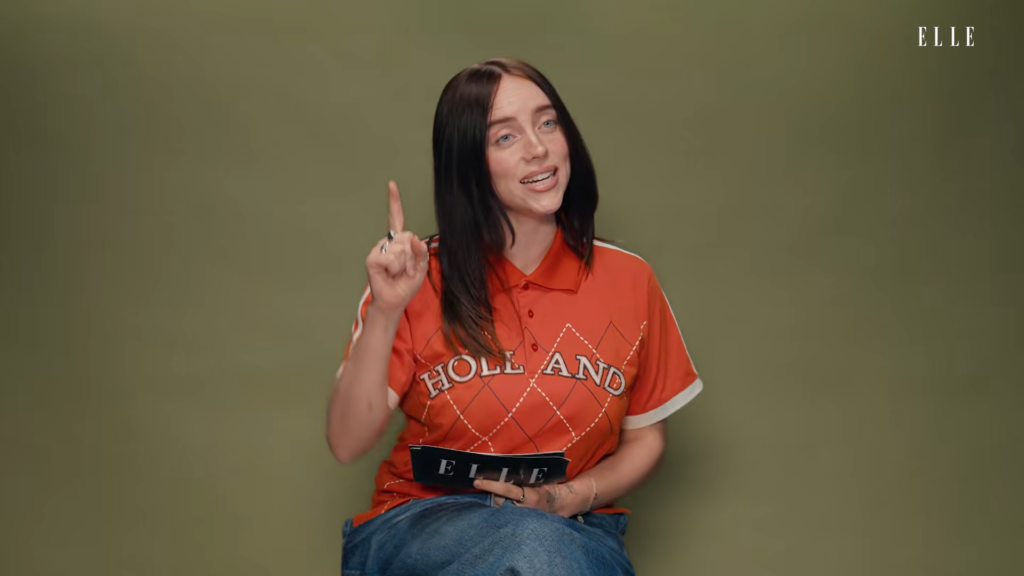 Woman with black hair in an orange polo sits against an olive background, raising her right index finger and holding an ELLE magazine.