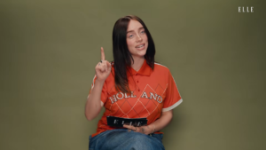 Woman with black hair in an orange polo sits against an olive background, raising her right index finger and holding an ELLE magazine.