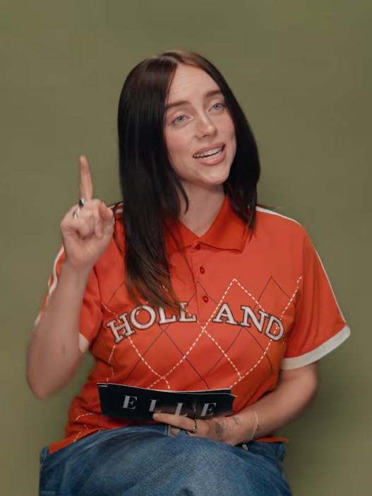 Woman with black hair in an orange polo sits against an olive background, raising her right index finger and holding an ELLE magazine.