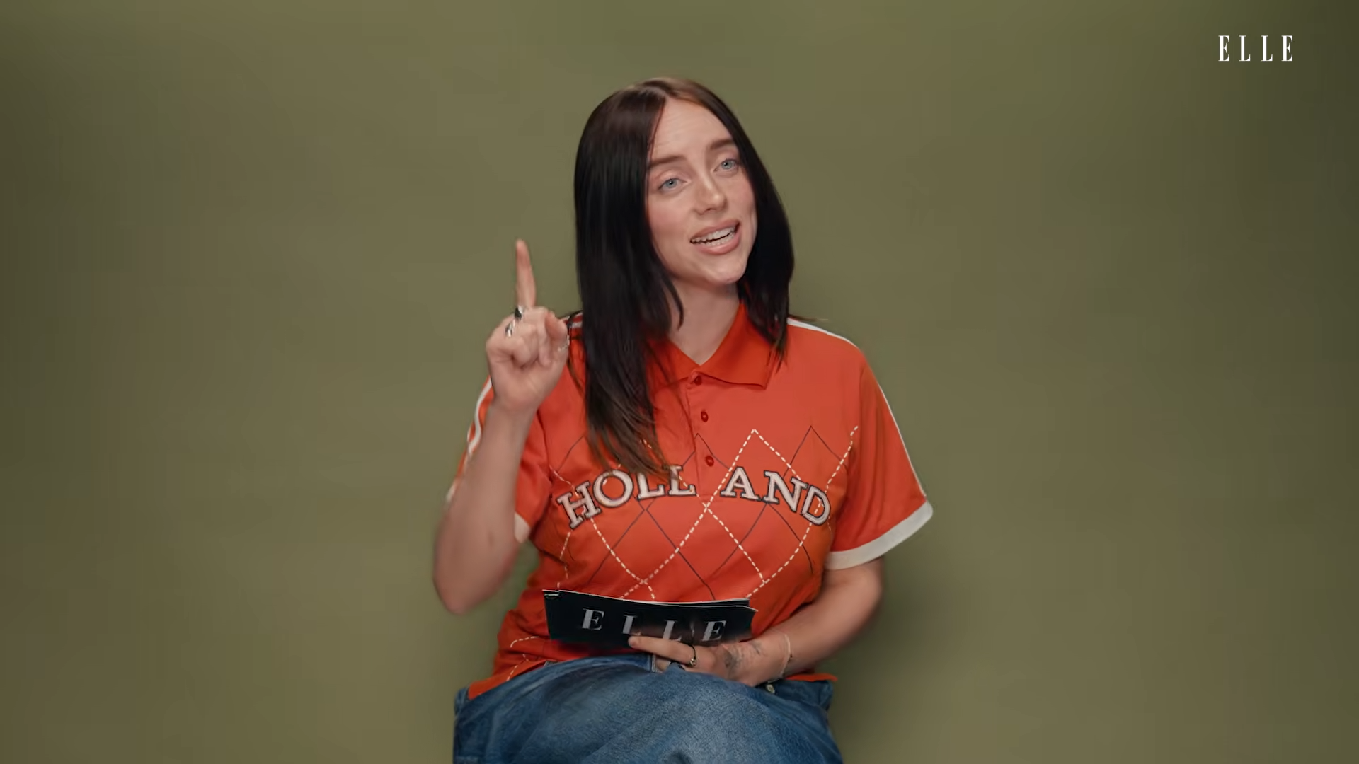Woman with black hair in an orange polo sits against an olive background, raising her right index finger and holding an ELLE magazine.