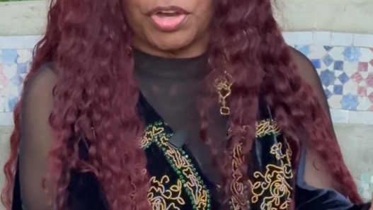 A woman with long maroon hair wearing a navy and gold embroidered outfit sits outdoors, speaking with hands raised as she gestures.