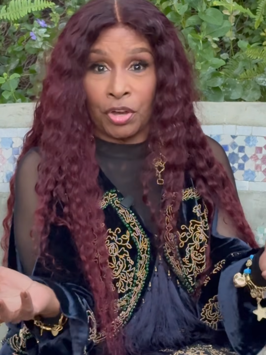 A woman with long maroon hair wearing a navy and gold embroidered outfit sits outdoors, speaking with hands raised as she gestures.