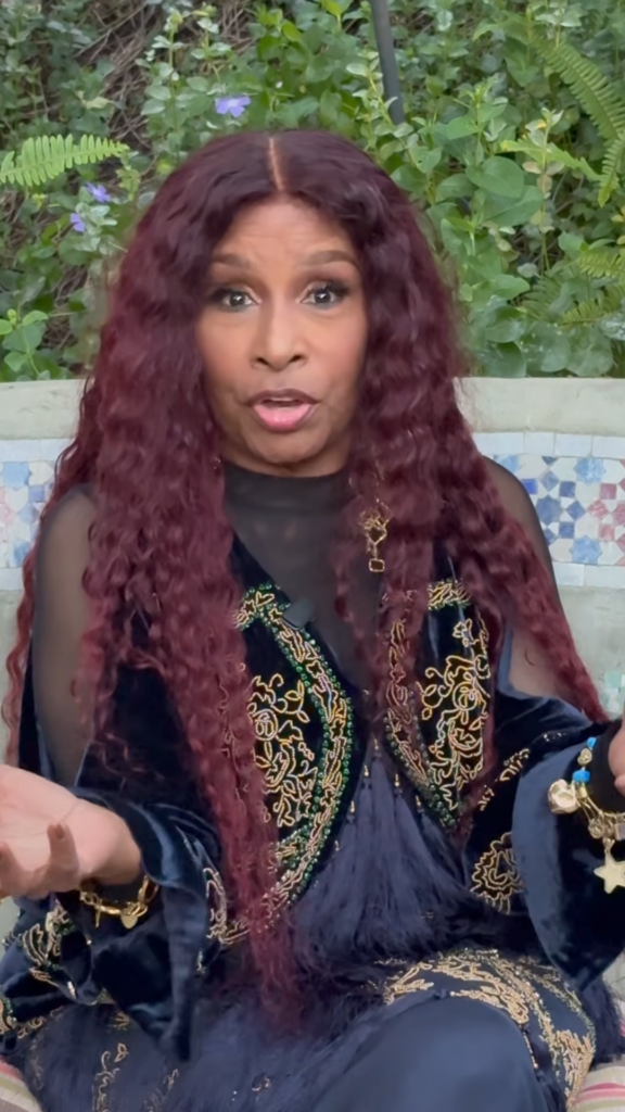 A woman with long maroon hair wearing a navy and gold embroidered outfit sits outdoors, speaking with hands raised as she gestures.