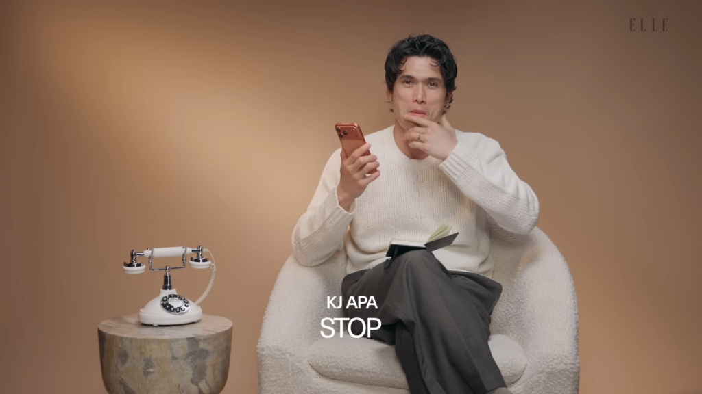 Man in a white knit sweater sits in a fluffy chair, holding a rose‑gold smartphone and a notebook, with a vintage rotary phone on a wooden stump to the left, ELLE logo in the top-right.