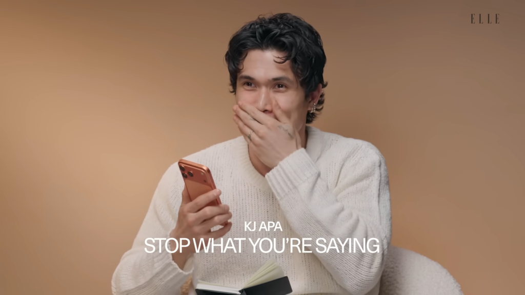 Man in a white sweater smiles with his hand over his mouth while holding a red smartphone, against a tan backdrop (Elle logo in corner).