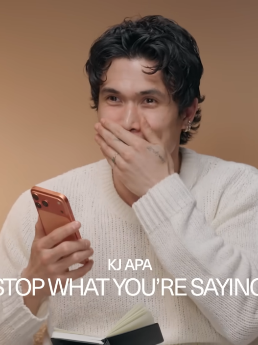 Man in a white sweater smiles with his hand over his mouth while holding a red smartphone, against a tan backdrop (Elle logo in corner).