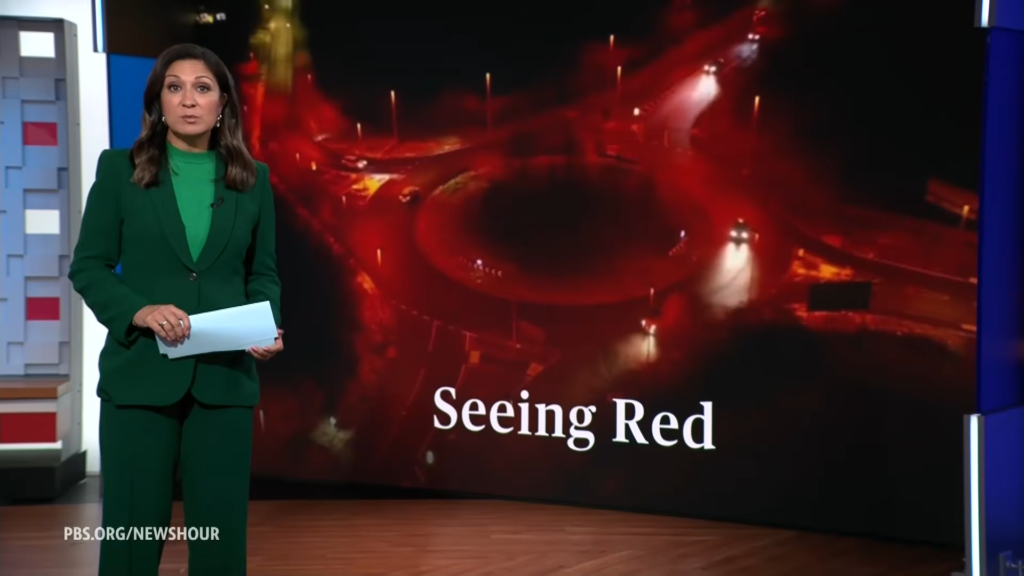 PBS NewsHour anchor in a green suit stands at the desk while a large screen behind shows red-toned graphics and the caption 'Seeing Red'.