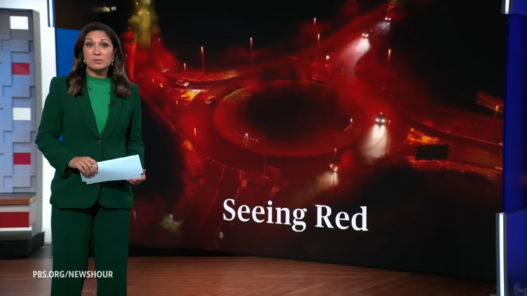 PBS NewsHour anchor in a green suit stands at the desk while a large screen behind shows red-toned graphics and the caption 'Seeing Red'.