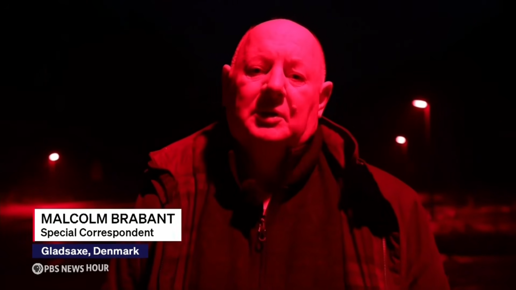 Close-up of a man speaking in red lighting, with a lower-left caption reading 'MALCOLM BRABANT, Special Correspondent' and PBS NewsHour logo nearby.