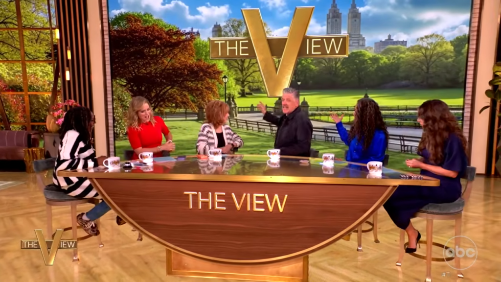 Six hosts sit around a curved wooden desk on The View set, mugs in front, mid-conversation.
