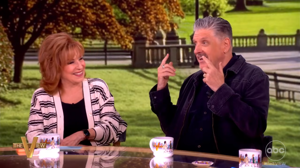 Two hosts sit at a glass desk on a daytime talk show, smiling and gesturing as they chat, with a park-like backdrop behind them.