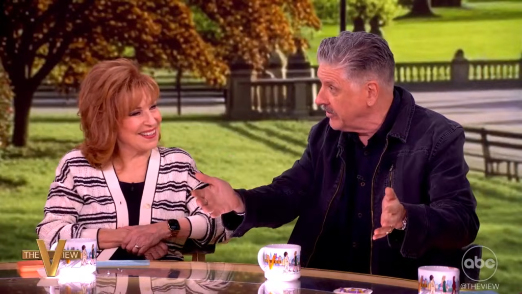 Two talk show hosts sit at a glass table outdoors, smiling as they shake hands with a park backdrop behind them, including trees and benches.