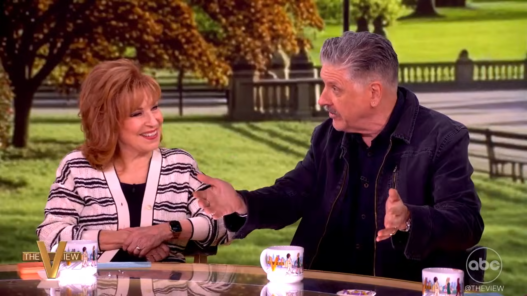 Two talk show hosts sit at a glass table outdoors, smiling as they shake hands with a park backdrop behind them, including trees and benches.