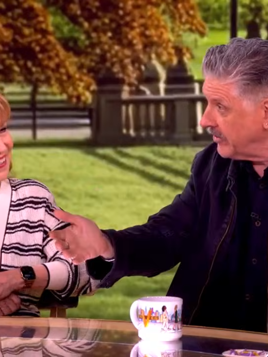 Two talk show hosts sit at a glass table outdoors, smiling as they shake hands with a park backdrop behind them, including trees and benches.