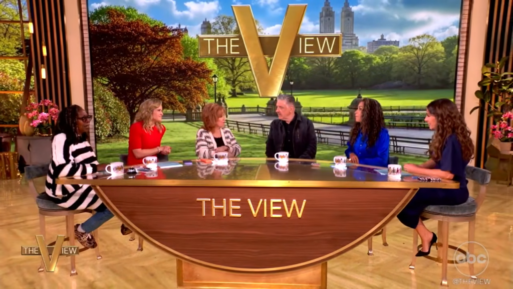 Six hosts sit around a curved wooden table on The View set, with a park backdrop and large THE VIEW sign behind them.