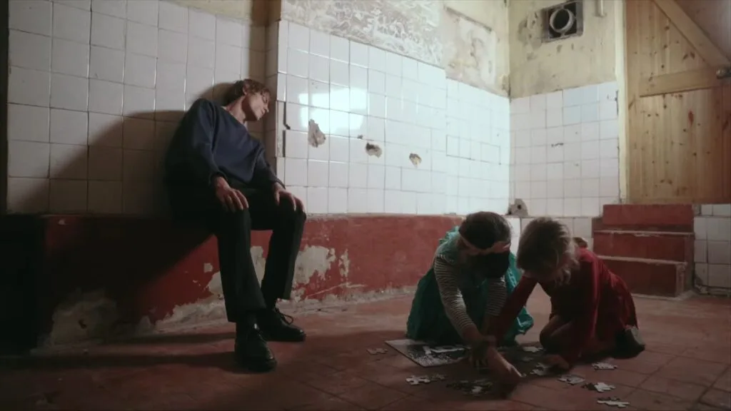 Man sitting on a red bench against a tiled wall, gazing downward as two children on the floor work on a puzzle in an abandoned-looking room.