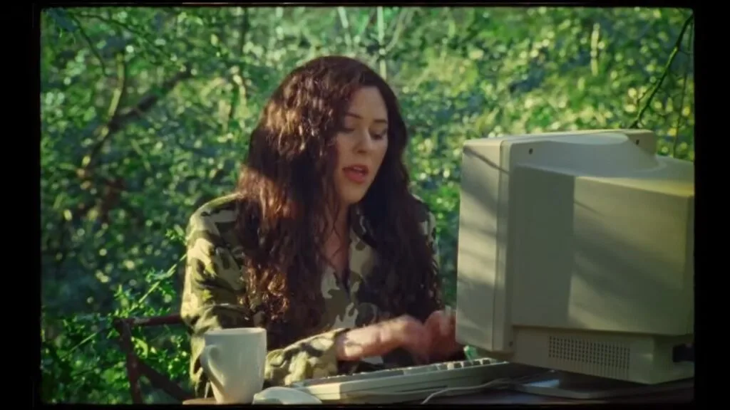 Woman in a camouflage shirt typing on an old desktop computer at an outdoor table among green trees, with a mug nearby.