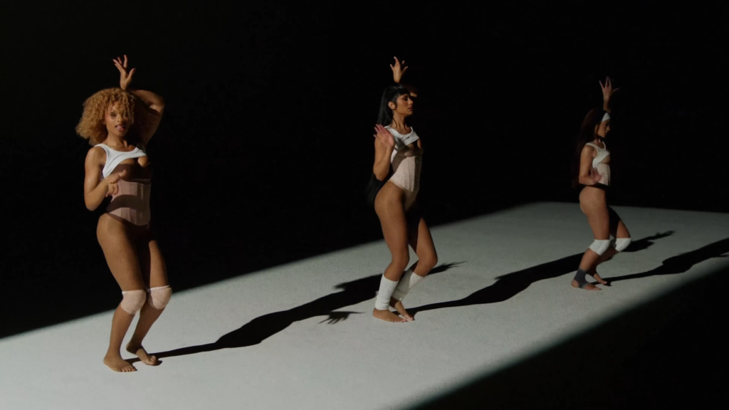 Three female dancers in light-colored leotards perform on a lit stage, long shadows across the floor.