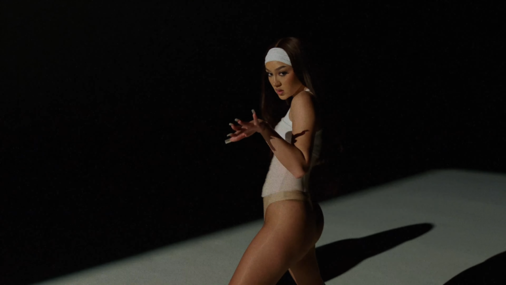 Female dancer in a white sleeveless outfit and headband poses on a lit stage against a dark background.