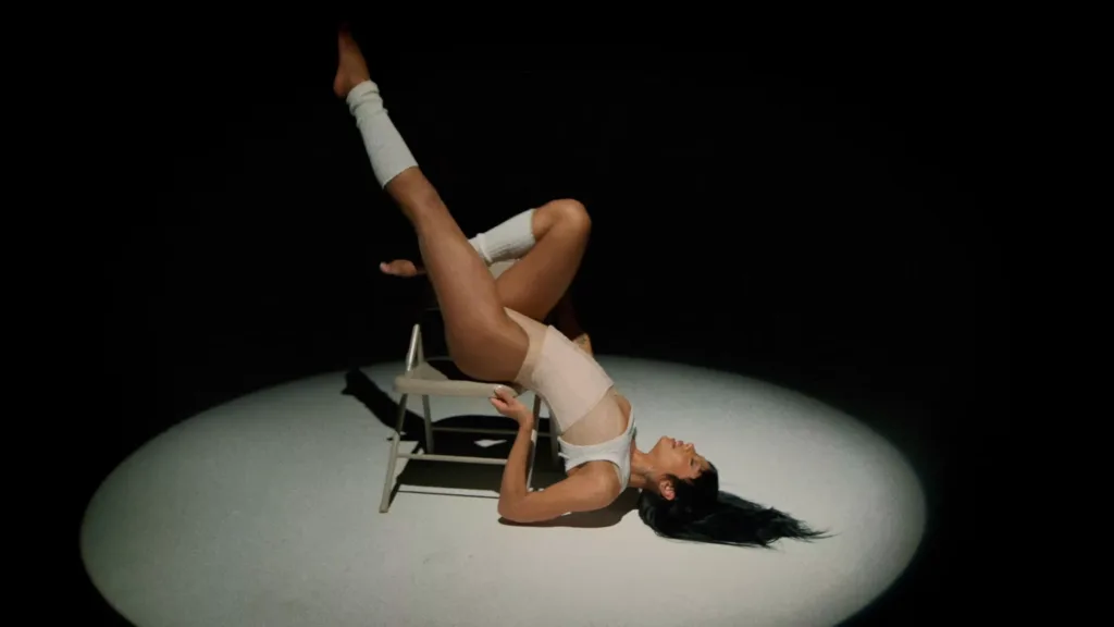 Dancer in white athletic wear balances on a small chair under a spotlight, legs extended overhead with white wraps on calves and shins.