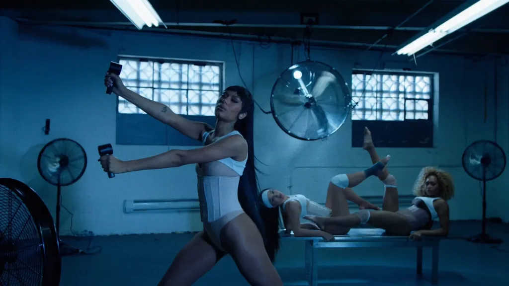 Three women in a blue-lit industrial studio exercise: one in front stands and extends arms with dumbbells while two others stretch on a table behind her.