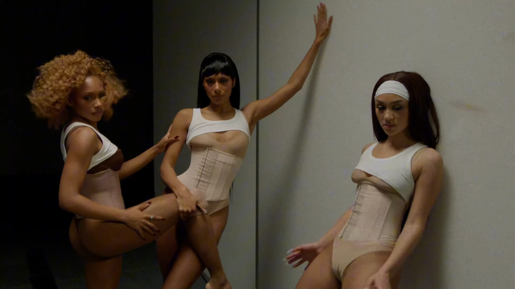Three women in nude shapewear and white crop tops pose against a wall in a studio, each showing different leg and arm positions.