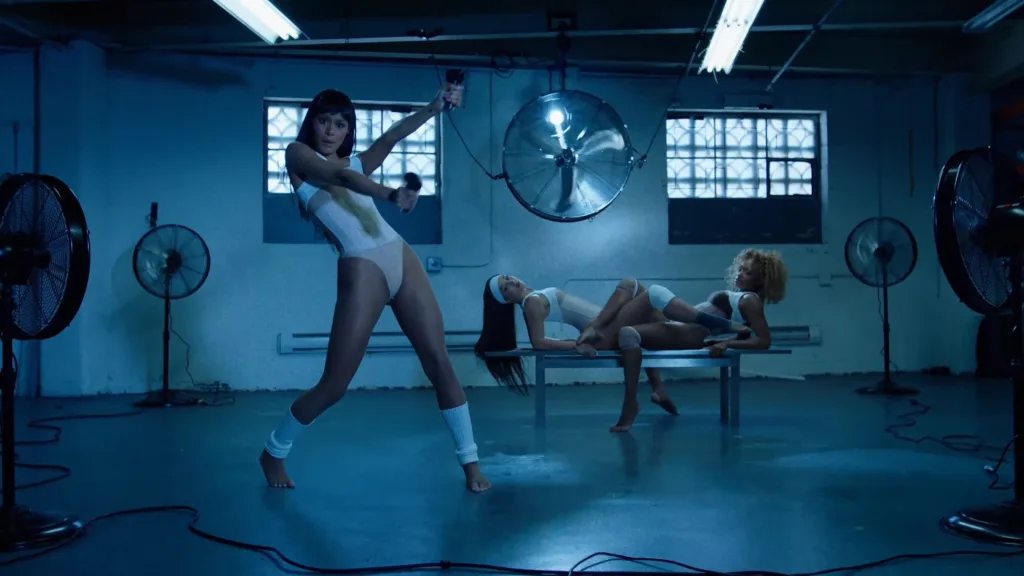 Three dancers in a blue-lit studio; one in a white leotard performs with arms extended, while two others pose on a bench in the background.
