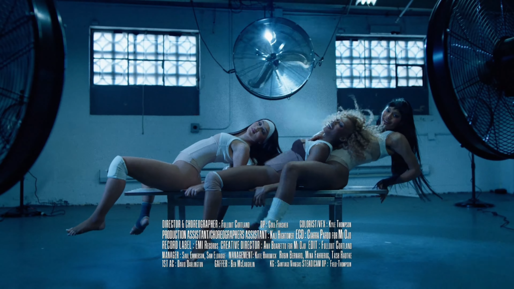 Three women in white athletic outfits perform back-to-back splits on a metal table in a blue-lit industrial studio, large fans flanking the scene with credits text at the bottom.
