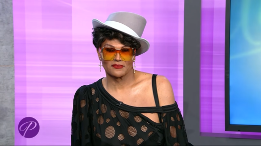 Person wearing a gray fedora, orange-tinted sunglasses, and a black mesh off-shoulder top on a purple TV studio background.