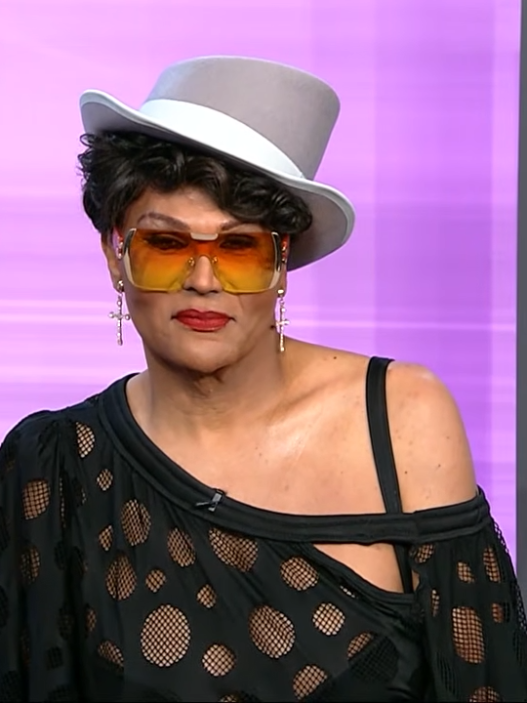 Person wearing a gray fedora, orange-tinted sunglasses, and a black mesh off-shoulder top on a purple TV studio background.