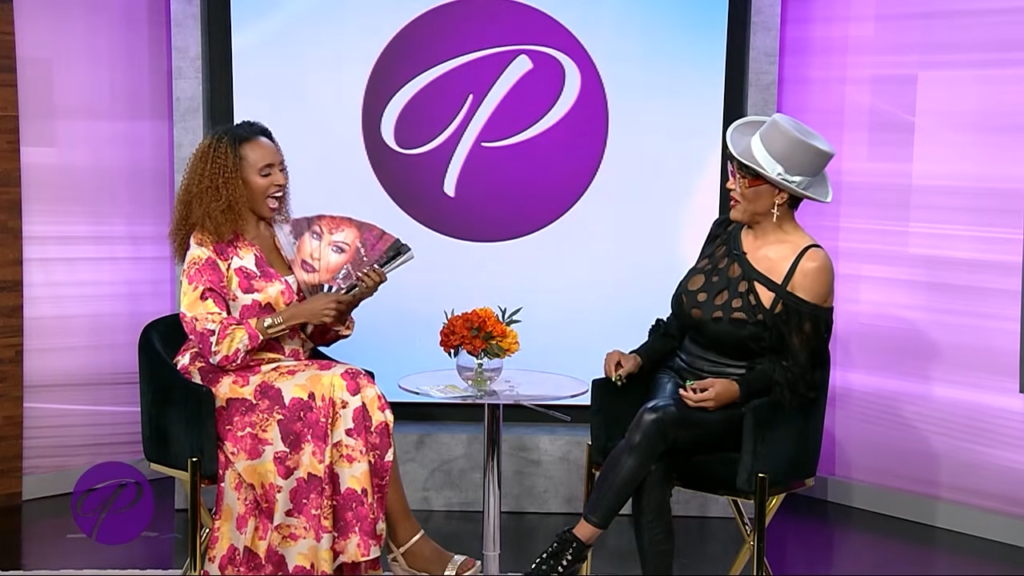 Two women seated opposite each other in a colorful TV studio, one reading fan magazines while the other listens in a black cutout top and hat.