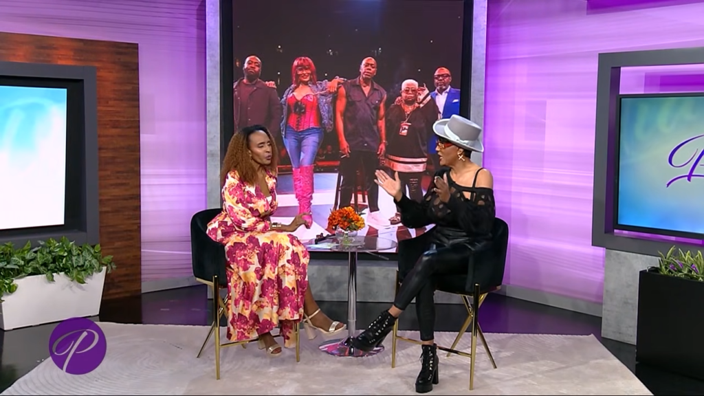 Two women sit facing each other on a purple-lit talk-show set; one in a floral dress, the other in black with a gray hat, with orange flowers on a glass table between them and a large group photo on screen behind.