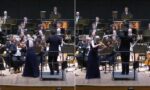 Conductor leads a full orchestra on stage as a violinist in a dark gown plays at the front.