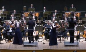 Conductor leads a full orchestra on stage as a violinist in a dark gown plays at the front.
