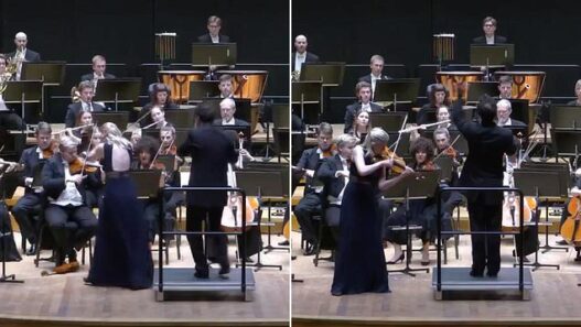 Conductor leads a full orchestra on stage as a violinist in a dark gown plays at the front.