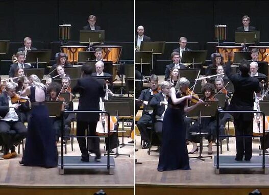 Conductor leads a full orchestra on stage as a violinist in a dark gown plays at the front.