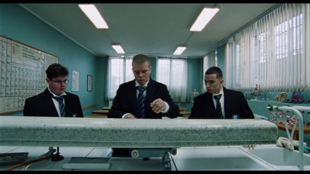 Three students in suits stand at a lab bench in a teal science classroom, examining a large metal instrument.