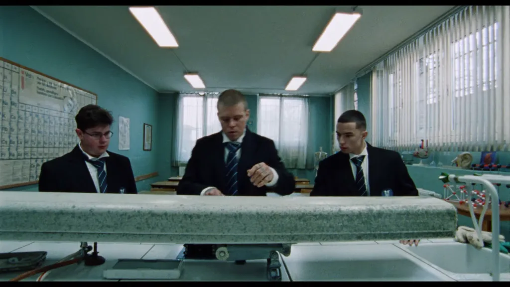 Three students in suits stand at a lab bench in a teal science classroom, examining a large metal instrument.