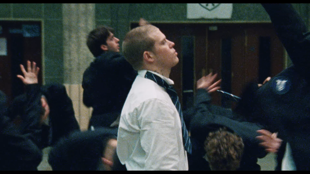 Man in a white shirt and striped tie stands among a crowd, looking upward with a serious expression.