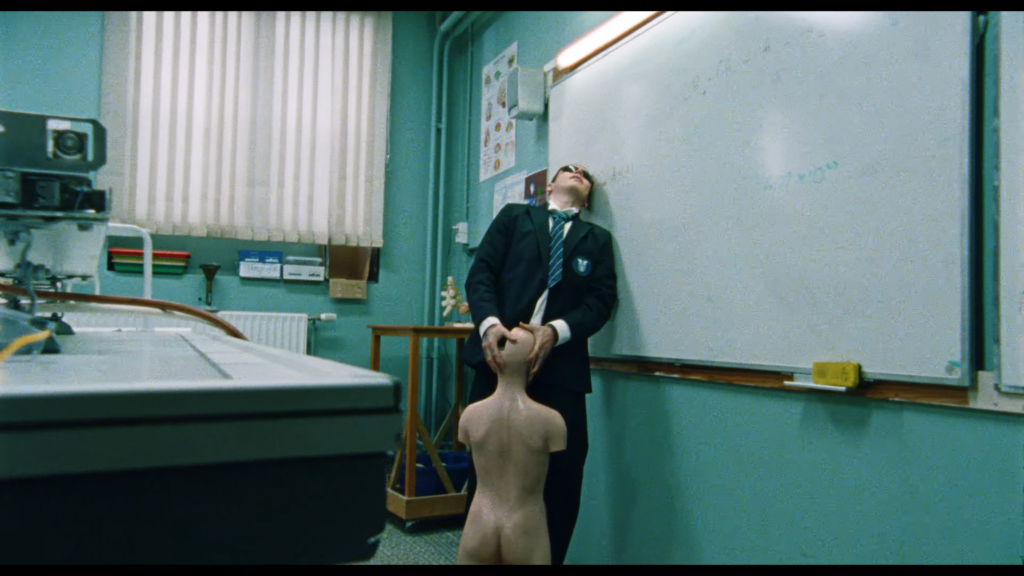 Man in a blazer and tie leaning against a classroom wall with a head tilted back; a nude mannequin torso is in the foreground.