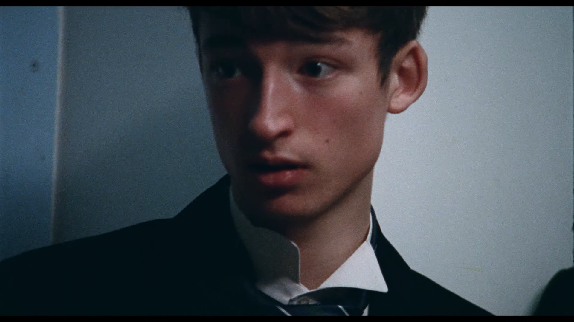 Close-up of a young man in a dark suit looking to the left, against a neutral background.