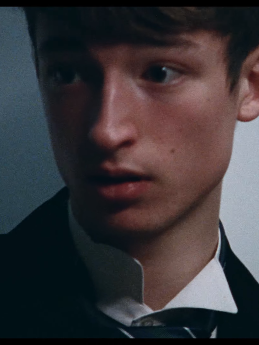 Close-up of a young man in a dark suit looking to the left, against a neutral background.